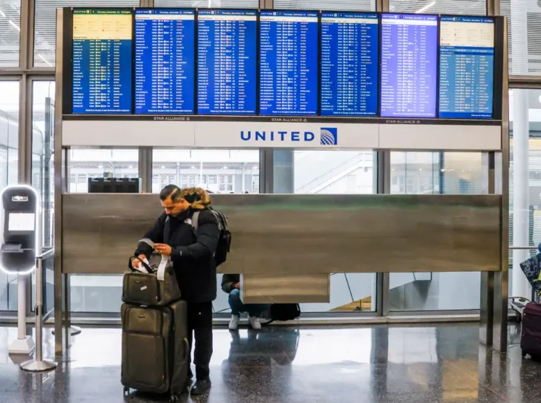 Un hombre junto a un tablero de horarios de vuelos mientras los pasajeros se registran para sus vuelos tratando de viajar ant